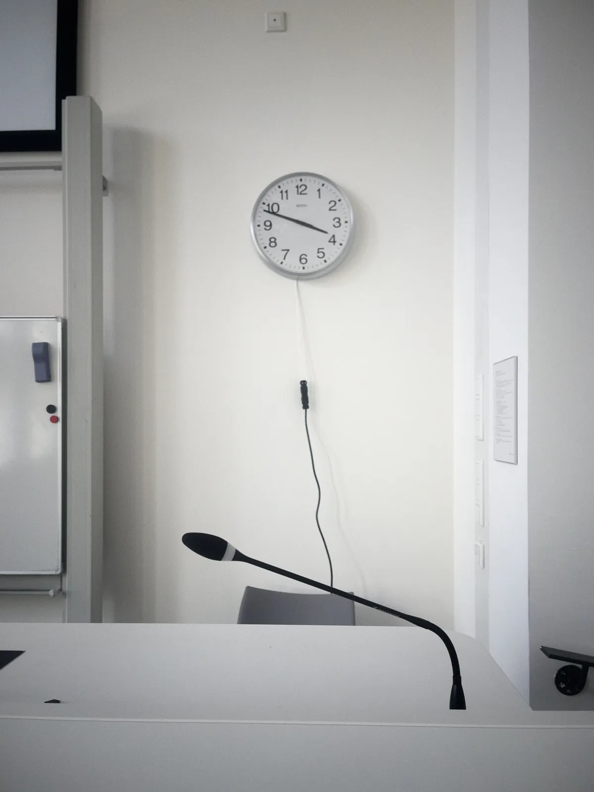 A small part of a whiteboard, a microphone on a stand in the foreground, a part of a chair, and a simple modernist wall clock on a white wall. It points at 12 minutes to 3.