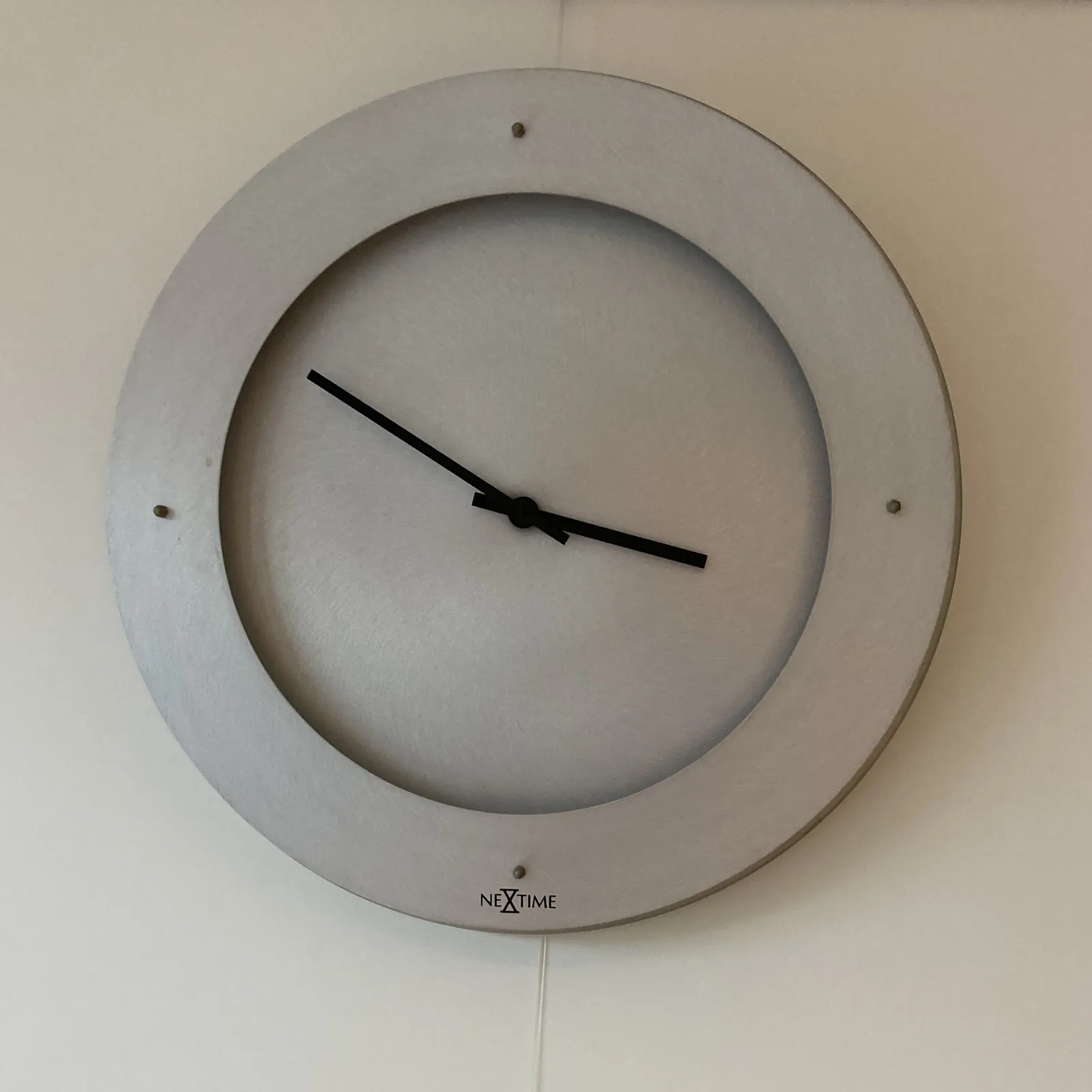 A rather minimalist wall clock. It consists of a wider ring, floating over a circle. Both are made from the same, gray material. On the ring there are four dots, or pins, indicating the four quarters of the clock. On the clock’s face the brand name Nextime is printed. The hands of the clock are black, and they point at 11 minutes to 4.