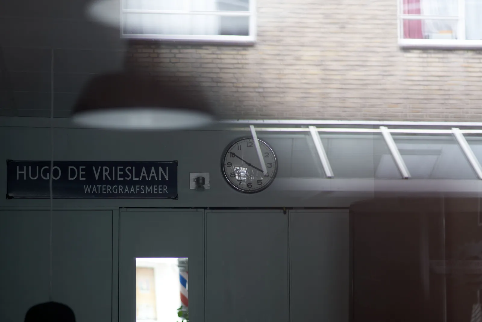 Looking into a barber shop. You can see a building reflected in the window. Confusingly there’s a street name sign inside the shop. It reads Hugo de Vrieslaan, Watergraafsmeer. There’s also a clock in the shop. It points at 10 to 4.