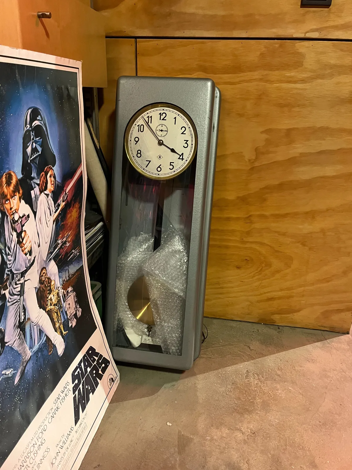 Next to a star wars poster, leaning against a wooden wall, stands a metal wall clock. It points at 7 minutes to 4