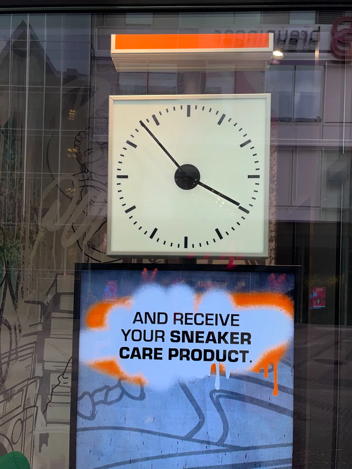 A square clock on top of a bright advertisement. It points at 7 minutes to 4. The advertisement reads And receive your sneaker care product.