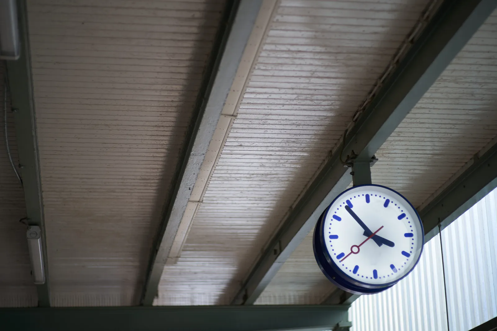 A clock, hanging from the ceiling in a train station. It points at 6 minutes to 4.