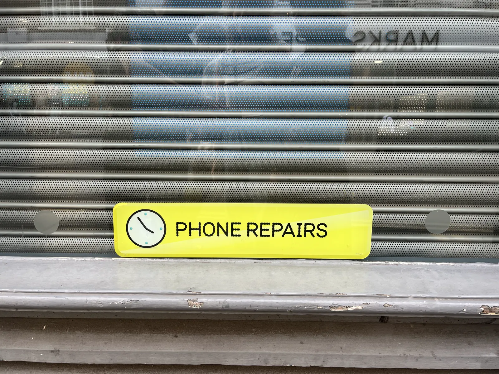 Rolled down, roll-down shutters with a yellow sticker on them which says Phone Repairs, next to an icon of a clock. This clock points at approximately 6 minutes to four.