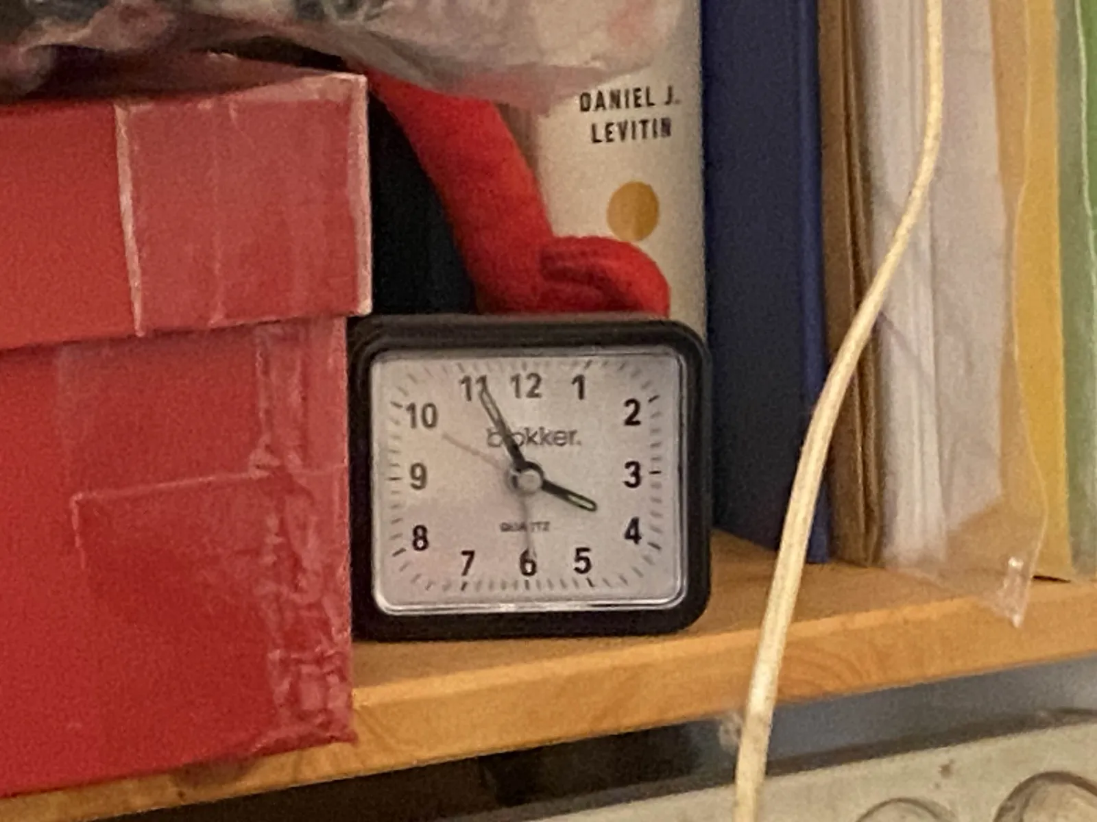 An alarm clock on a book shelf. It points at 4 minutes to 4.