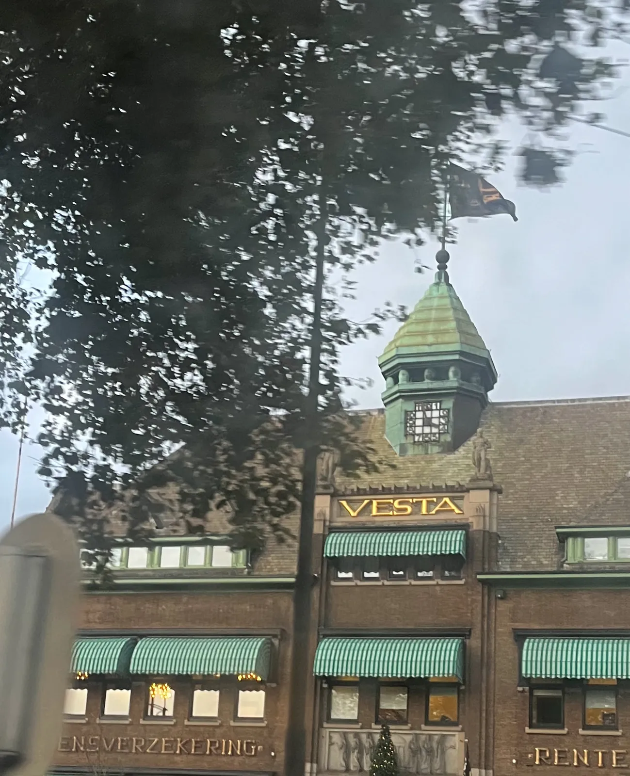 This picture seems to be taken from a moving vehicle. An older brick building with a small tower.It says Vesta in golden letters. There are white with green striped canopies in front of the windows. On the little tower there’s a clock, made from squares. It points at approximately 4 minutes to 4.