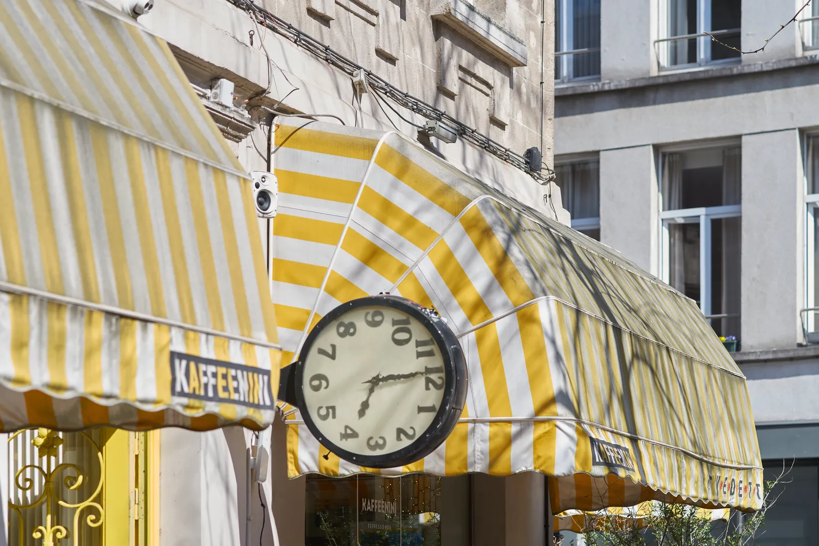 Two classic, striped sunblinds, yellow and white. Between them there’s a clock attached to the wall. It’s rotated 90 degrees, which of course makes it a bit surreal. If you turn your head it points at 1 minute to four.