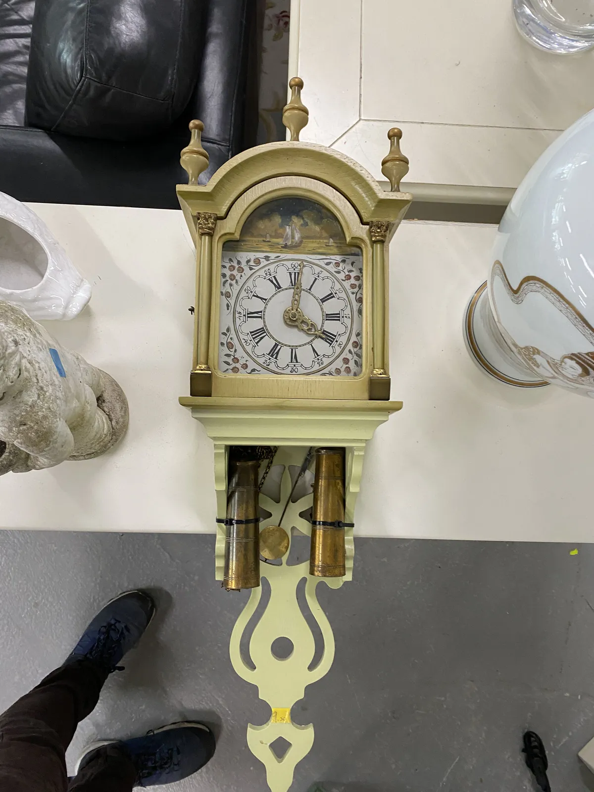 A classic wall clock laying on the floor, but pictured straight from above, so it seems like it hangs on the wall. The feet of the photographer show that this is not the case. The clock points at 1 minute past 4.