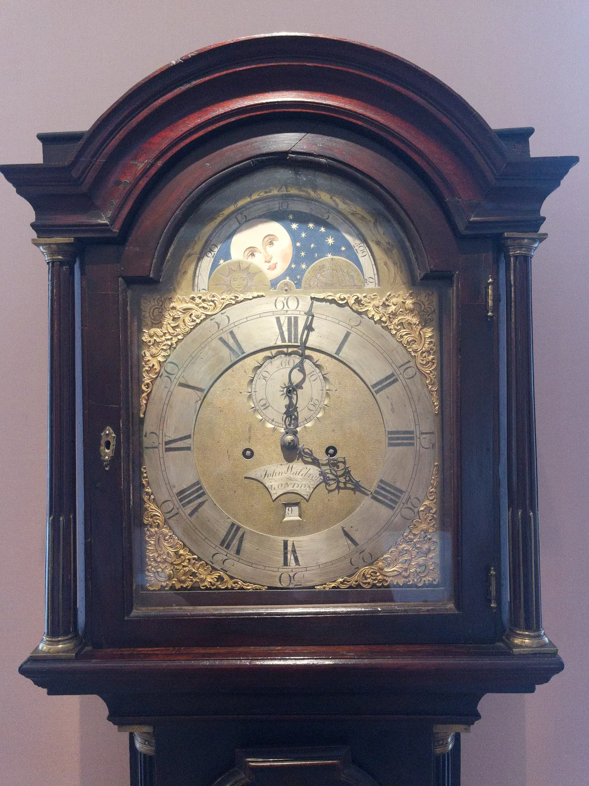 The top of a large, old, wooden standing clock. It has mechanics to show the day of the month, to show the time, of course, and mechanics to show the moon’s phases. The clock points at one minute past 4.