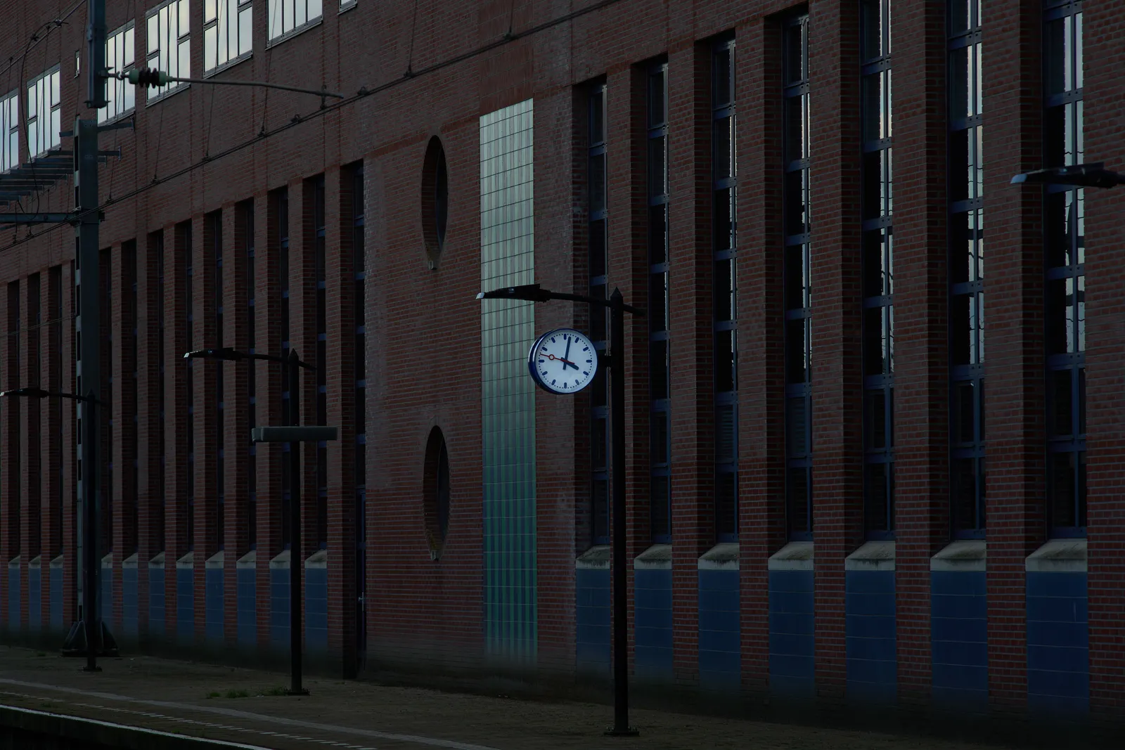 A high, long, brick wall with high, slim windows. There’s a clock attached to a pole, and it points at 2 minutes past 4.