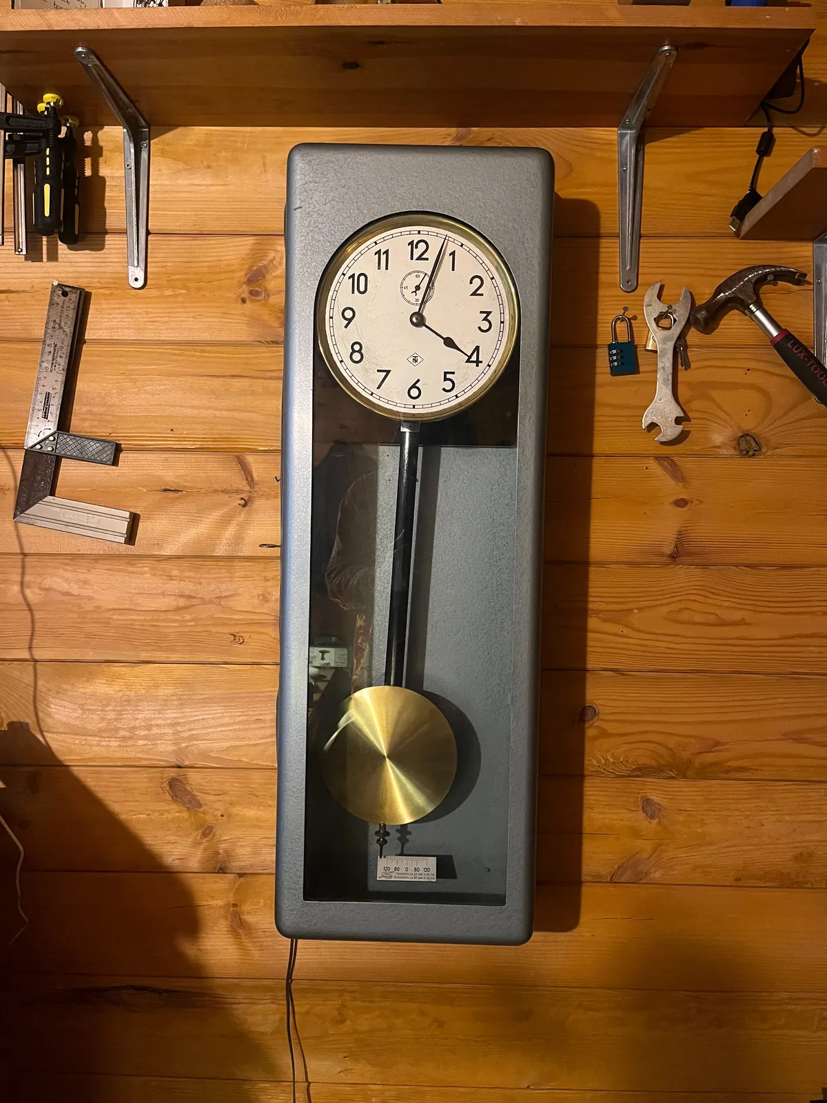 A grey metalic precision clock is hangin on a wall in a tool shed. It points at 3 minutes past 4