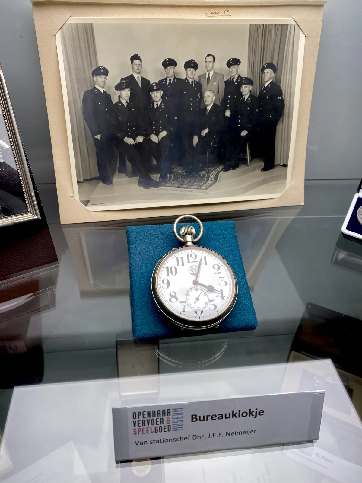 A little desk clock, on a little blue, satin pillow. It’s displayed in front of an old picture of a group of uniformed men, posing. The clock points at 3 minutes past 4.
