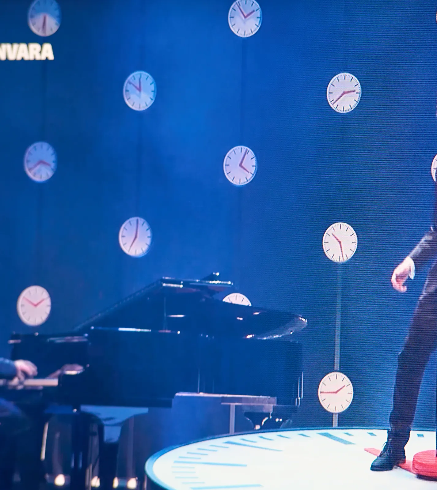 A picture taken from a television. On screen is a piano and part of a performer behind a microphone. The set is made from lots of clocks. Ten clocks are visible. They point at 31 minutes past six, 6 minutes to two, 9 minutes to 12, 22 minutes to 3, 21 minutes to 4, 4 minutes past 4, 7 o’clock, 27 minutes past 10, ten minutes to 2 and the last one points at a quarter to two. One of these clocks shows the right time.