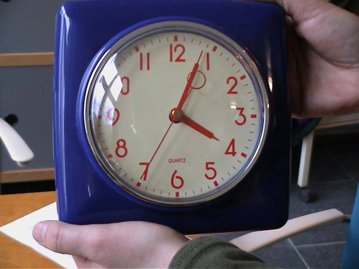 Somebody is holding a blue clock with orange numbers and arms in their hands. It points at four minutes past four.