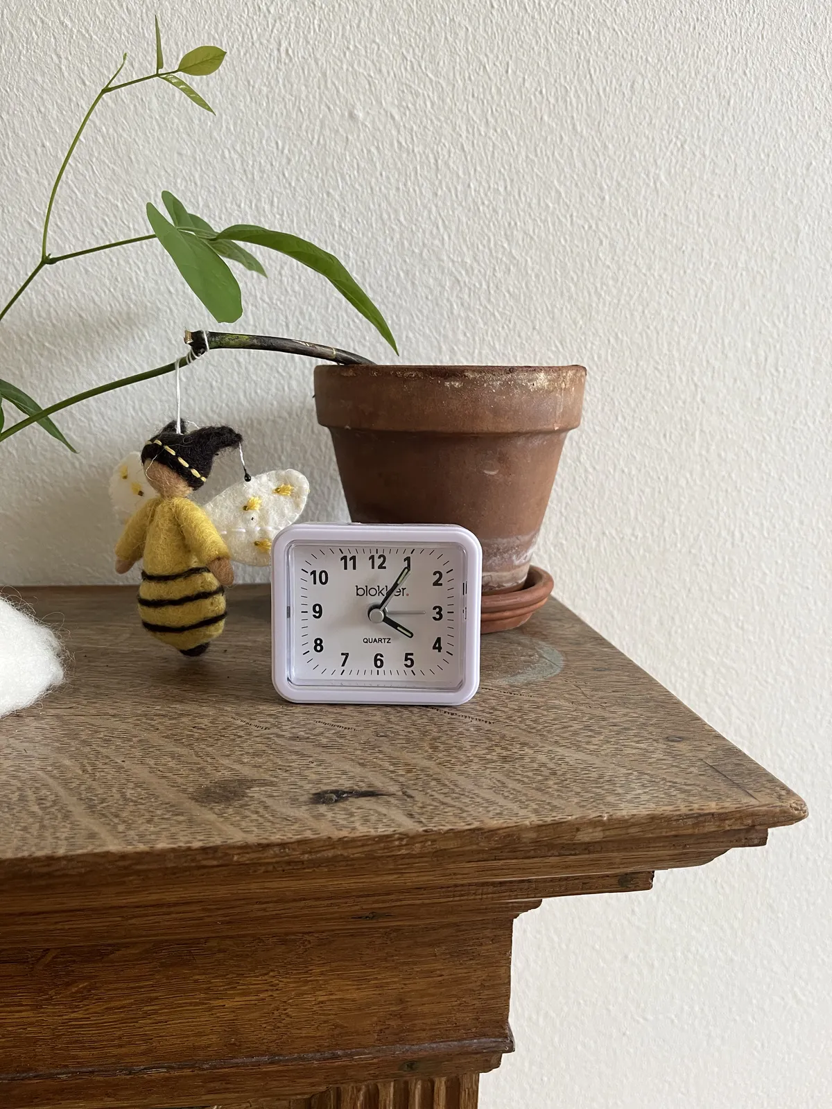 A woolen angle is hanging from a plant, next to a small, plastic alarm clock which points at 5 past four