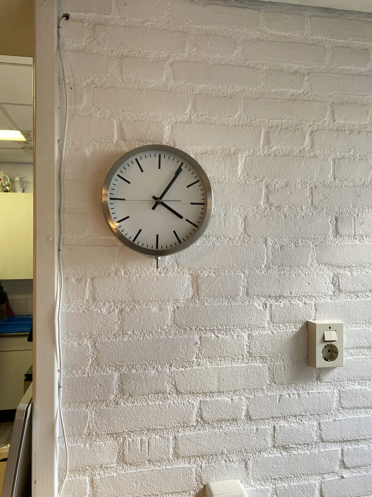 A white wall with a clock on it. It points at 5 minutes past 4. Next to the wall you can peek through a door. There’s not much you can see.  