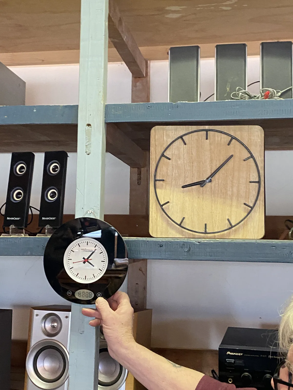 Speakers for sale, they are displayed on shelves. On one of these shelves there’s a clock. Someone is holding another clock in front of the shelves. The clock on the shelve points at 7 past 8, the other at 6 past 4.