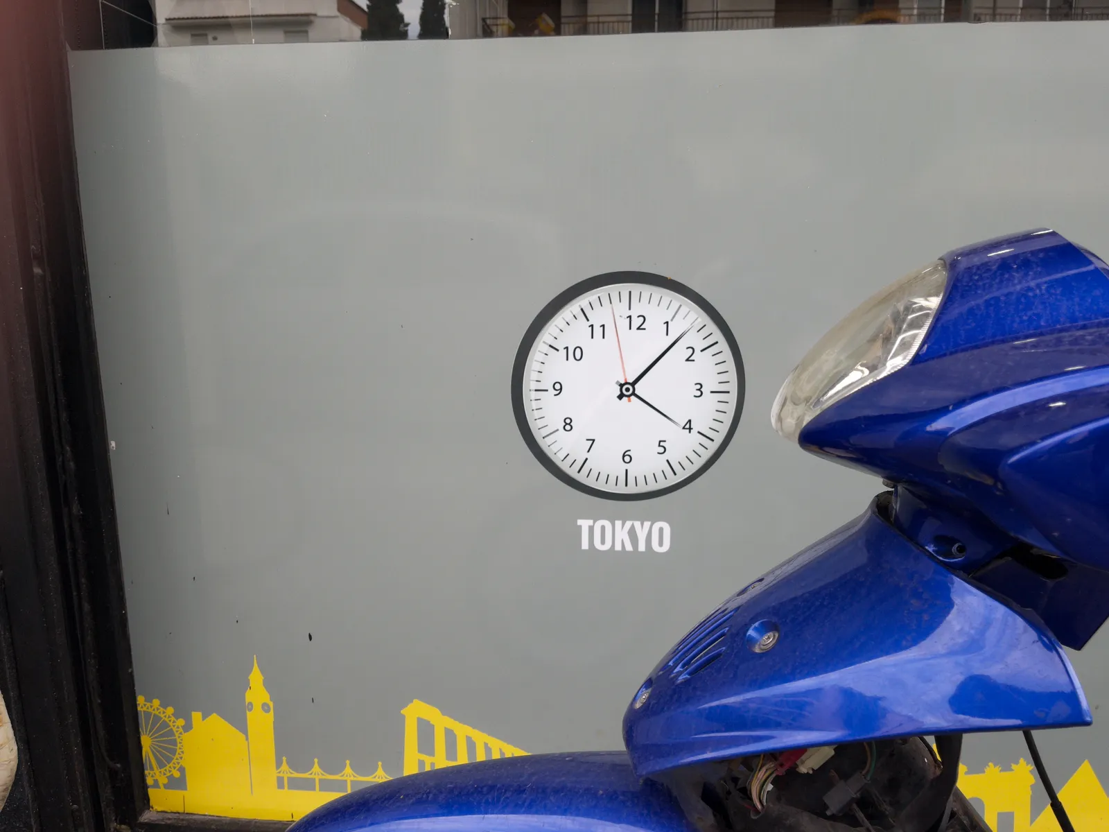 A blue scooter is parked in front a gray shopping window with a clock printed on it. It says Tokyo below it. The clock points at 7 past 4.