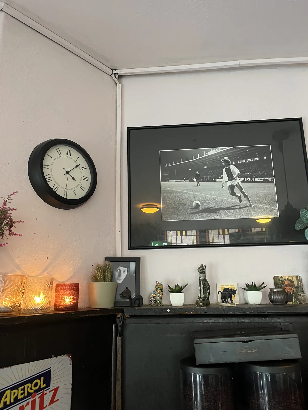 An old black and white picture on the wall. It’s a picture of a football (or soccer) player, playing the ball. There are candles, pictures of people, and statues of cats on a little shelf below the picture. Next to the picture, on the left, there’s a large wall clock which points at 7 minutes past four.