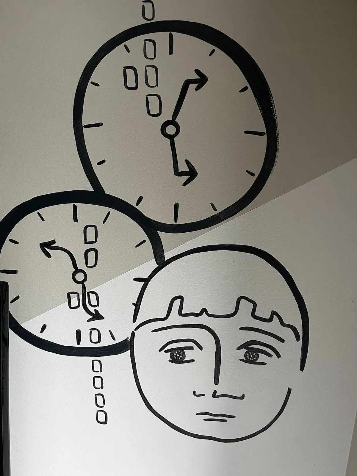 There’s a hand drawn face and two hand drawn clocks on this wall. All the hands of both clocks are wavy, or skewed, so assumptions have to be made. One of the clocks points at approximately 9 minutes past 4, the other at approximately 10 minutes to five. One of them is right.