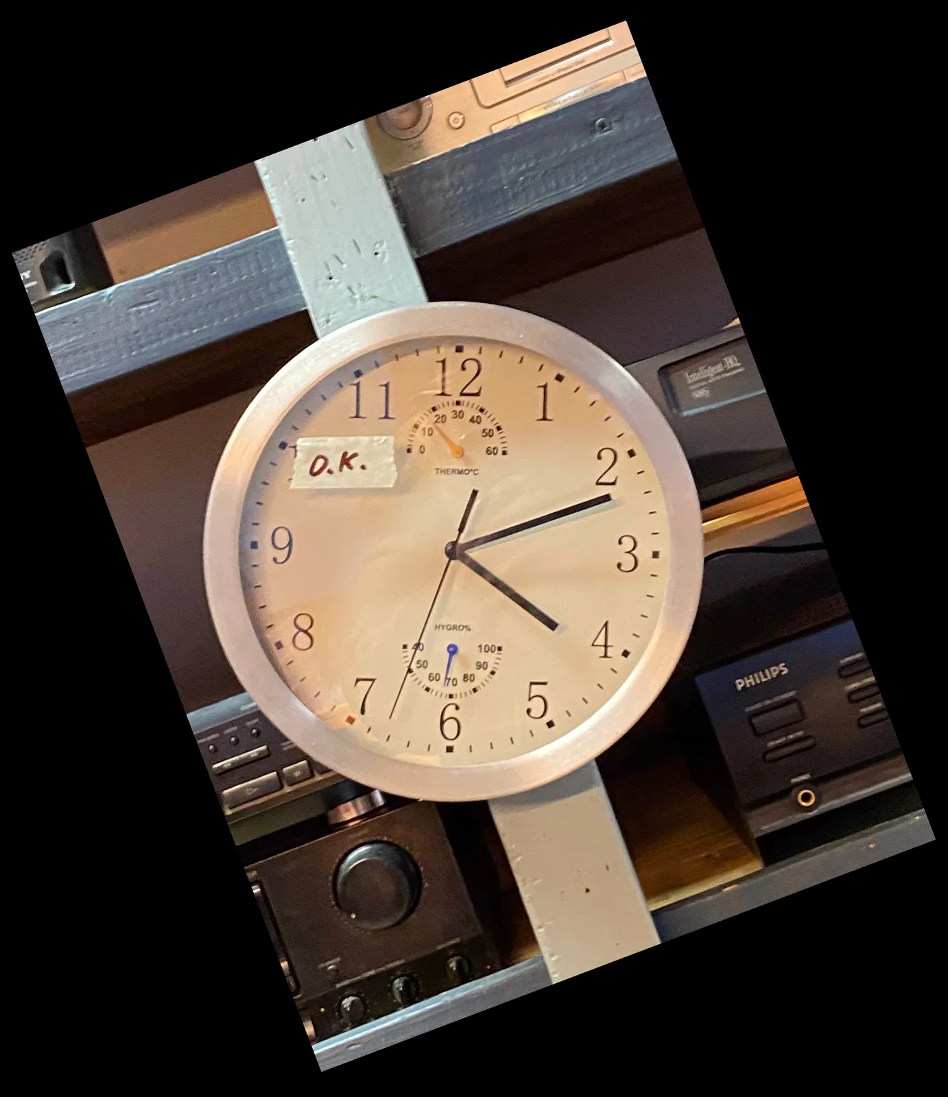 This clock is hanging crooked on a rack with audio equipment. The photographer skewed the image, so now the clock is straight, but the rack is slanted. A bit confusing in a nice way. The clock points at 12 past 4.