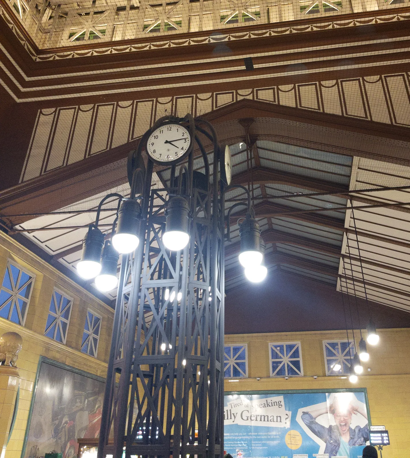 Looking up at a brown and white striped, high ceiling. There’s an enormous metal structure with a clock on it. It points at 13 minutes past 4.