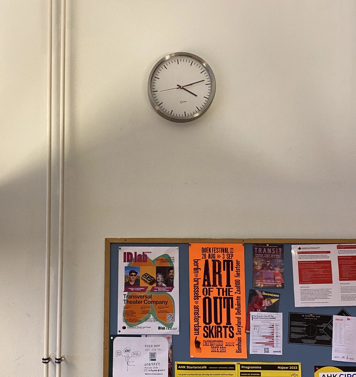 Posters on a bulletin board, above it there’s a clock. It points at 13 minutes past 4.