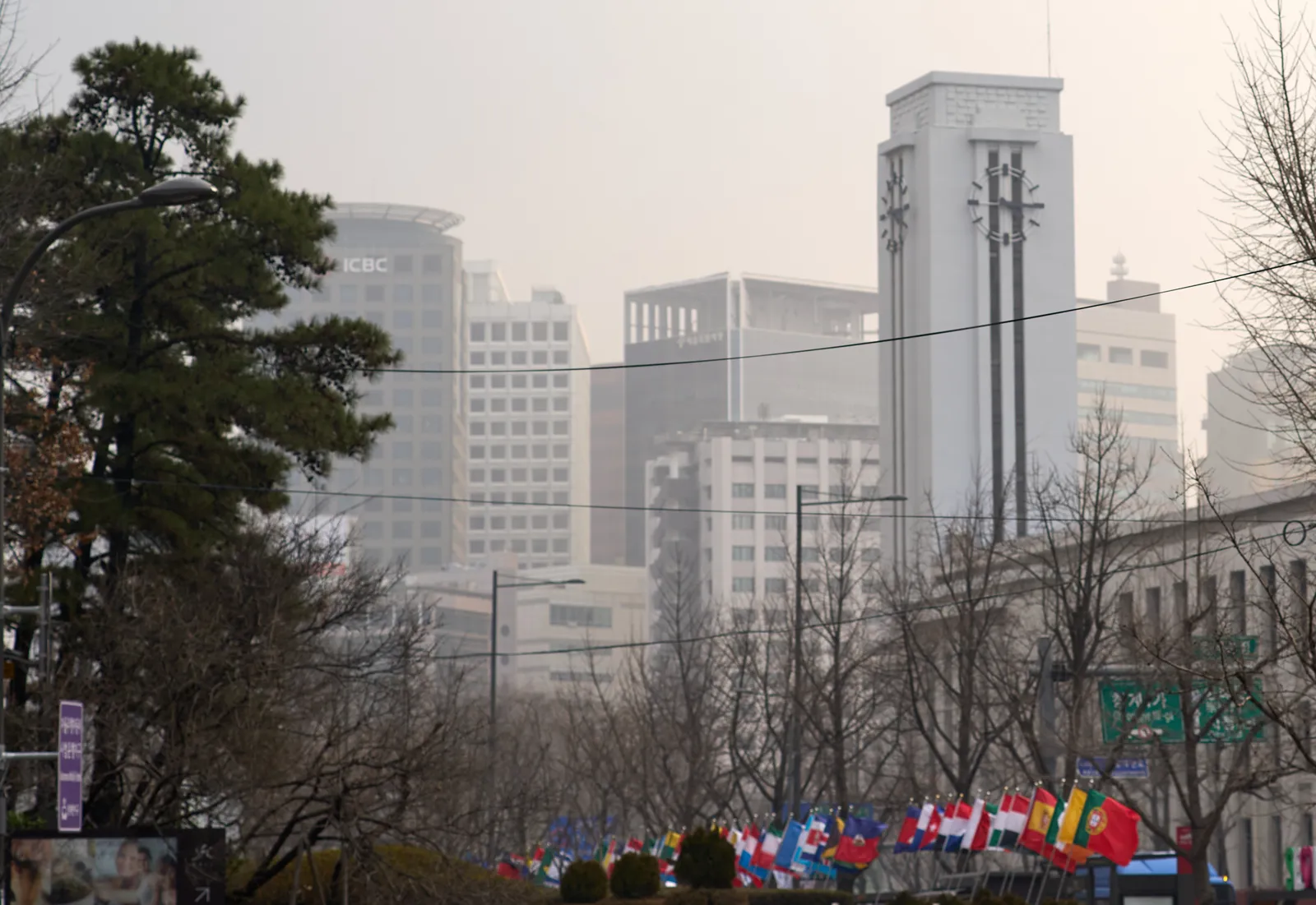 Many colourful flags, and some trees on a square. In the background, blurred by smog, some high buildings. In front of these buildings there’s high tower with a clock on it. It points at a quarter past 4.