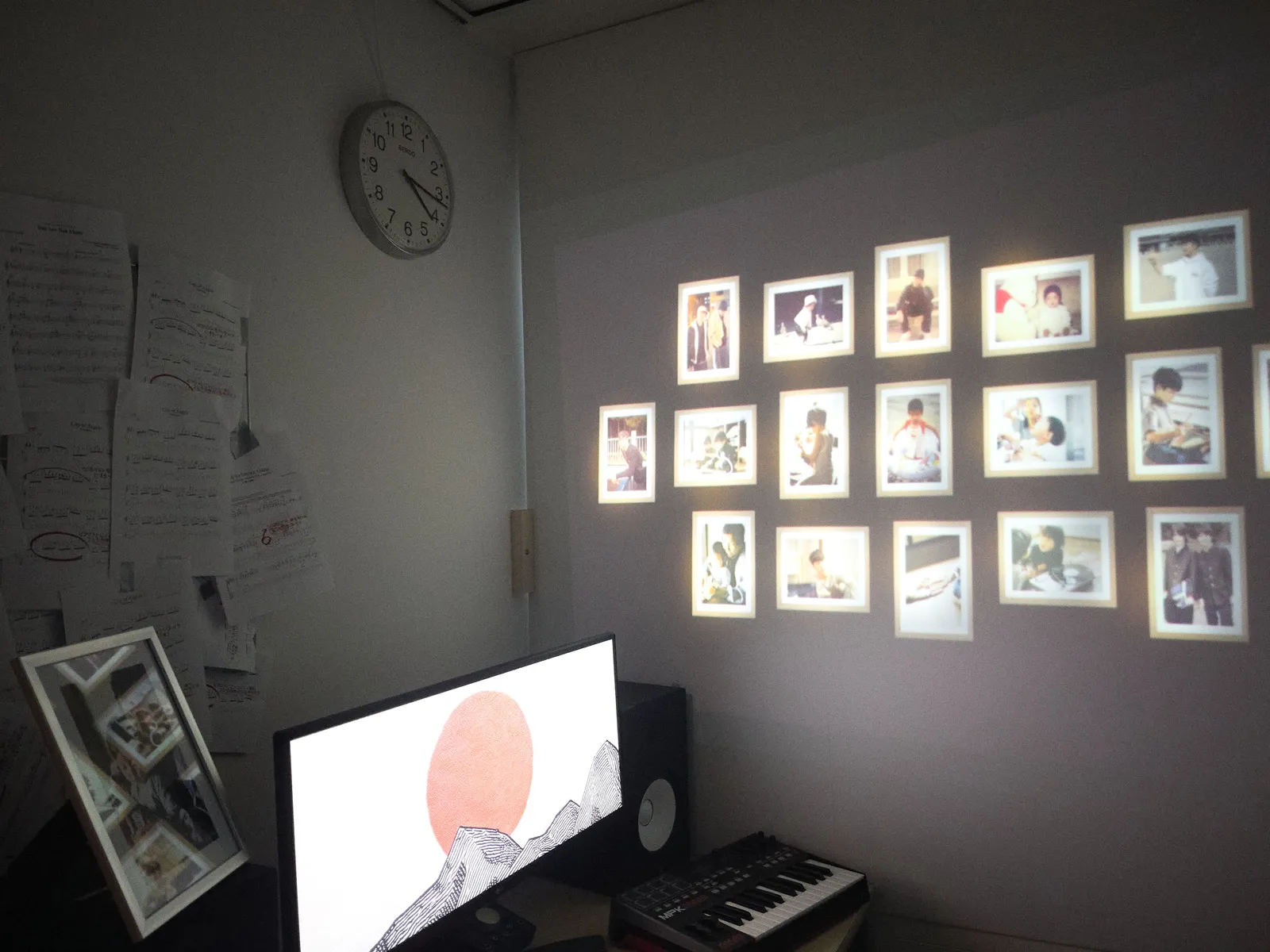 Papers pinned to a wall, images projected on another wall, a large computer screen with an image of a sun, a musical keyboard, and a clock. The clock points at 16 minutes past 4.