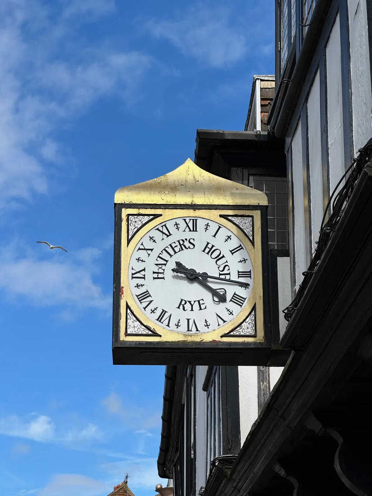 A clock attached to an old building. The clock has a golden triangle on top. A roof, or a hat, if you want to. On its face the words Hatter’s House Rye are printed, so both a roof and a hat make sense. The clock points at 17 minutes past 4.