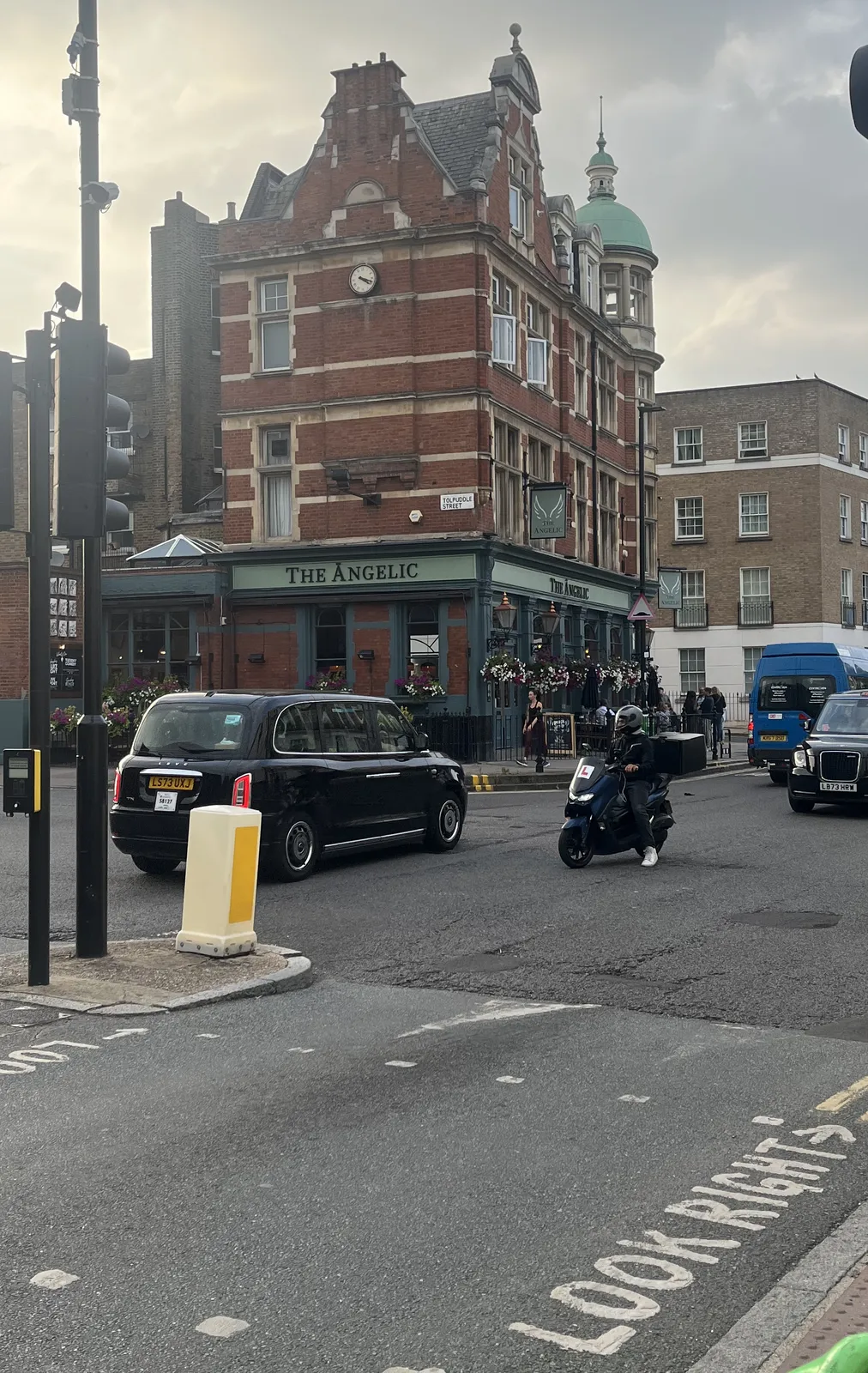 A crossroad. In the floor it says Look Right, with an arrow pointing rightwards. There are cars and motorcycles on the road, and in the background there’s a brick building with the name The Angelic on its facade. There’s also a clock, high on this building, which points at 17 minutes past four.