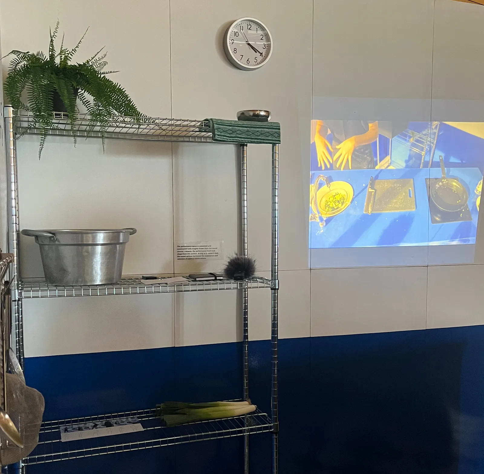 A projection of someone working in a kitchen on a wall. Next to this projection there’s a cupboard with kitchen things. There’s also a clock on the wall that points at 19 minutes past 4.