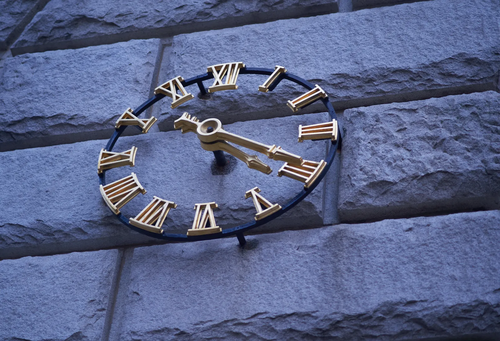 A closeup of a clock built into a monumental wall. The clock consists of golden roman numerals and two hands, gold as well. The clock points at 20 minutes past 4.