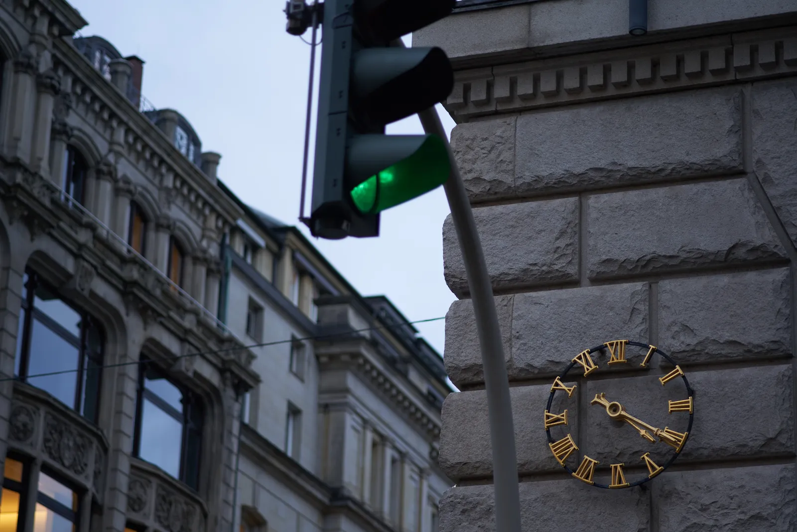 A green traffic light in the foreground, and an old monumental building in the back, which is a bit blurry. In focus there’s a clock with roman numerals built into a monumental wall. It points at 20 minutes past 4.