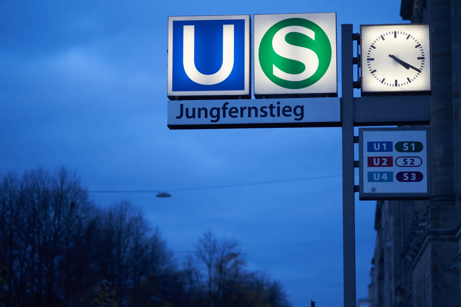 A lit sign that indicates that here, at Jungfernstieg station you can find both U-bahn trains and S-bahn trains. On an unlit sign it says the numbers of the trains: U1, U2 and U4, and S1, S2 and S3. There is a lit clock that points at 20 minutes past 4, so better hurry up a bit.