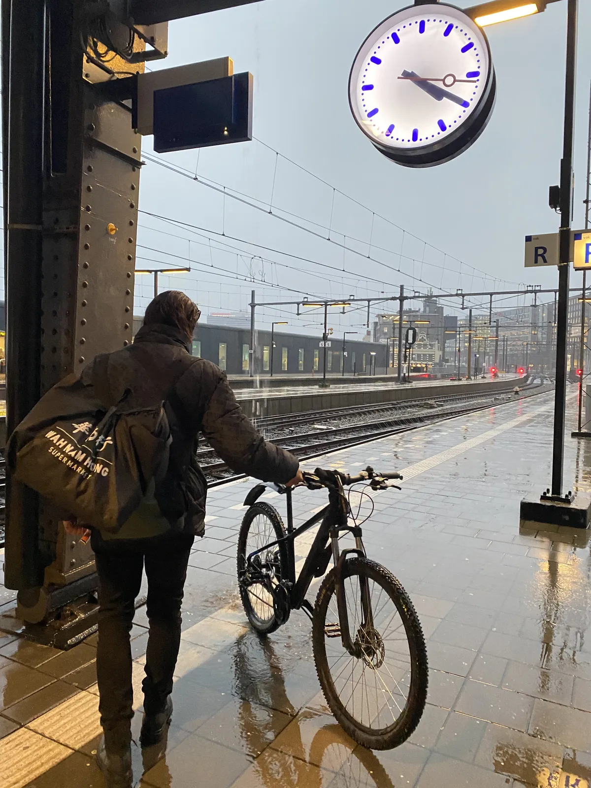 A person with a bike on the platform of a very, very rainy train station. The clock on the picture points at 20 past 4.