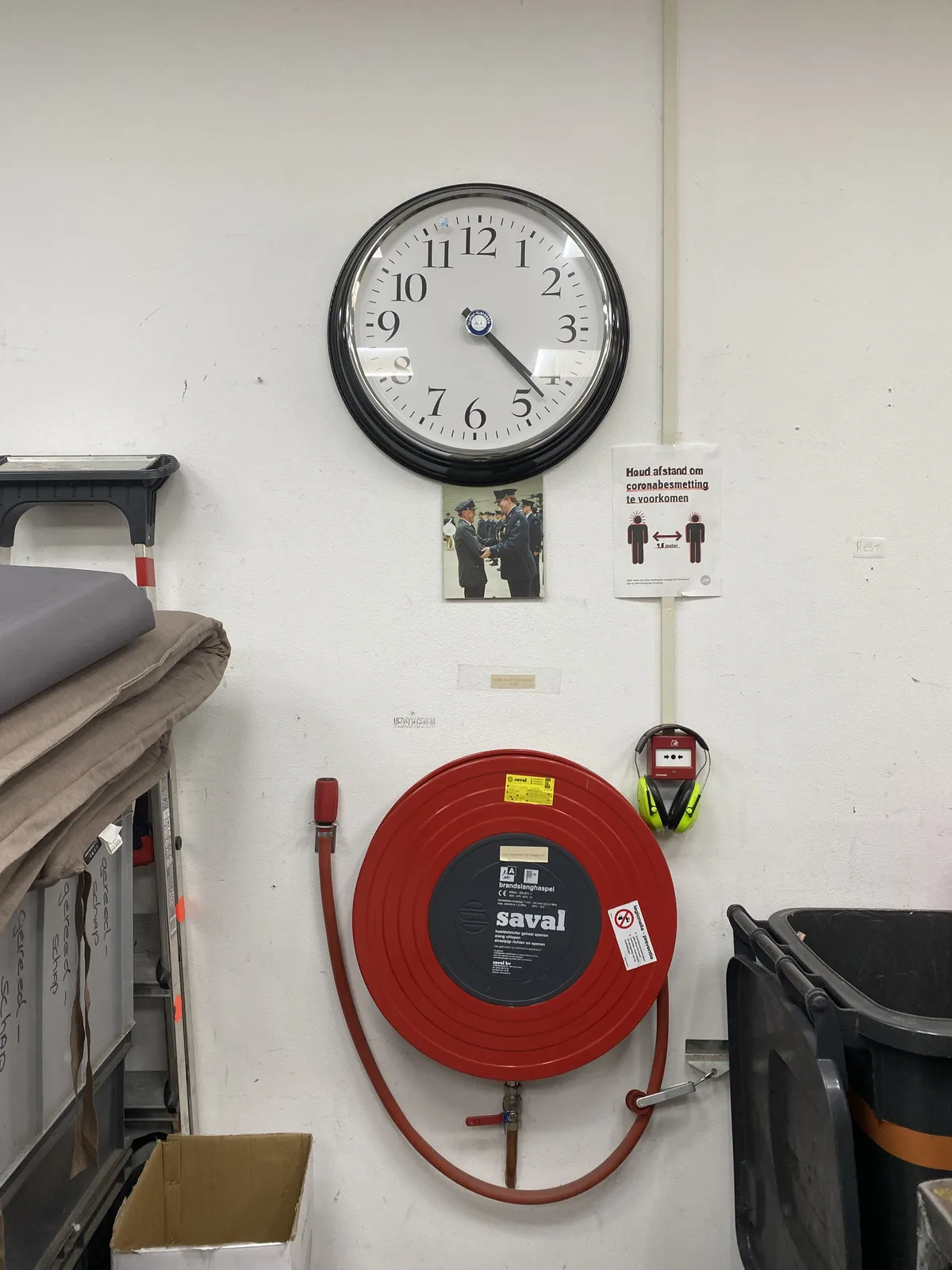 A firehose, earmuffs, a picture of the Dutch king shaking his grandfather’s hand, both in uniform, a sign that says to keep 1.5 meters distance, and a clock. On the clock it is 22 past 4.
