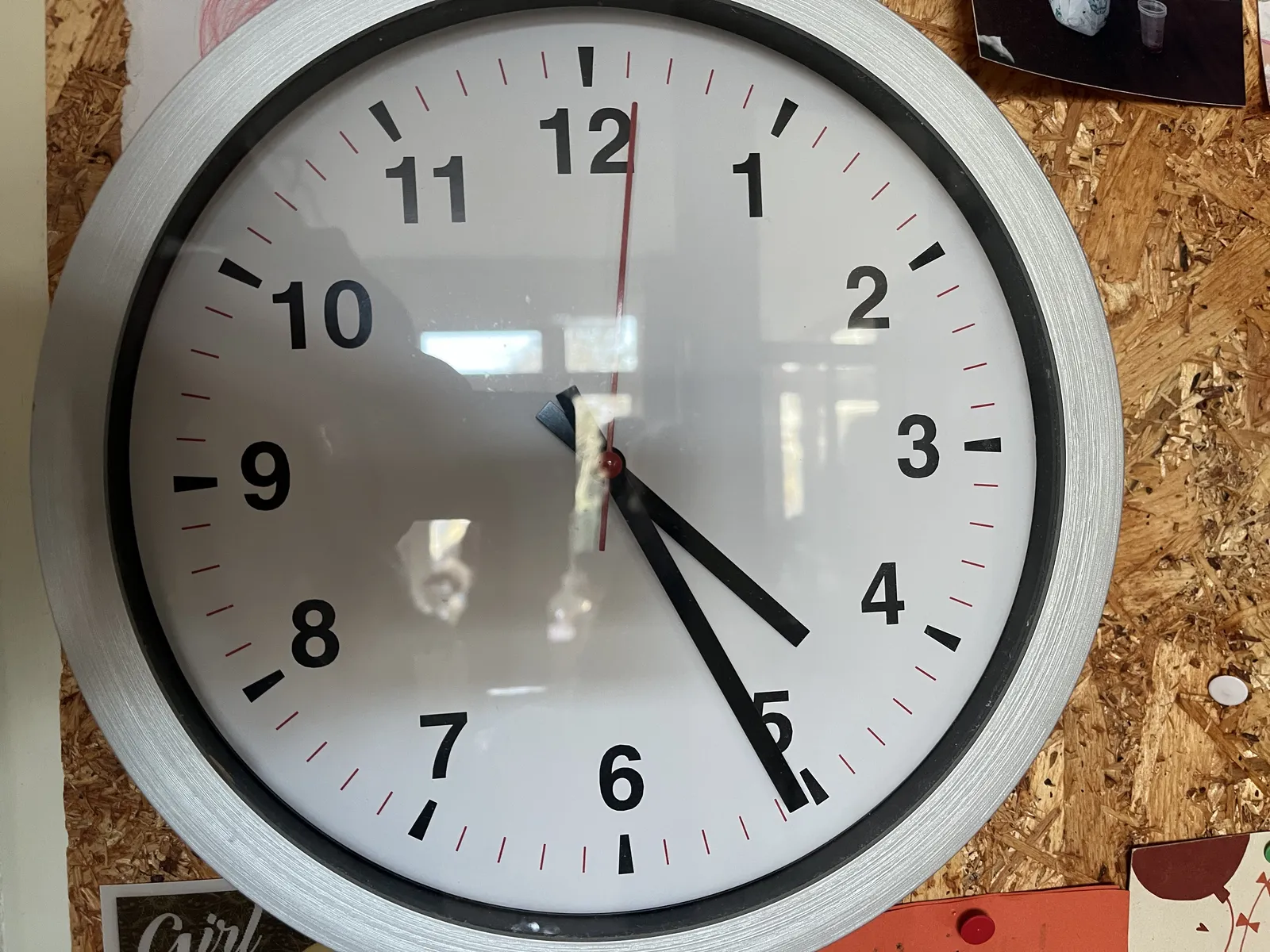 A closeup of a simple wall clock attached to a cork bulletin board. The clock points at 25 minutes past four.