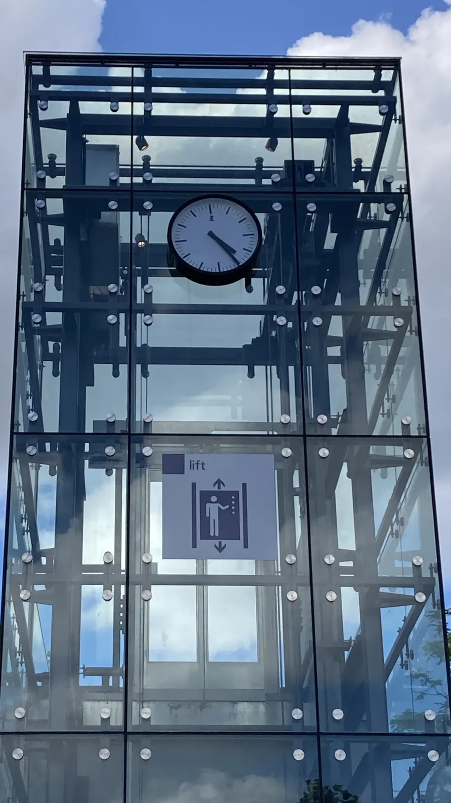 A glass stucture, probably for an elevator. On this structure there’s a clock that points at 25 minutes past 4.