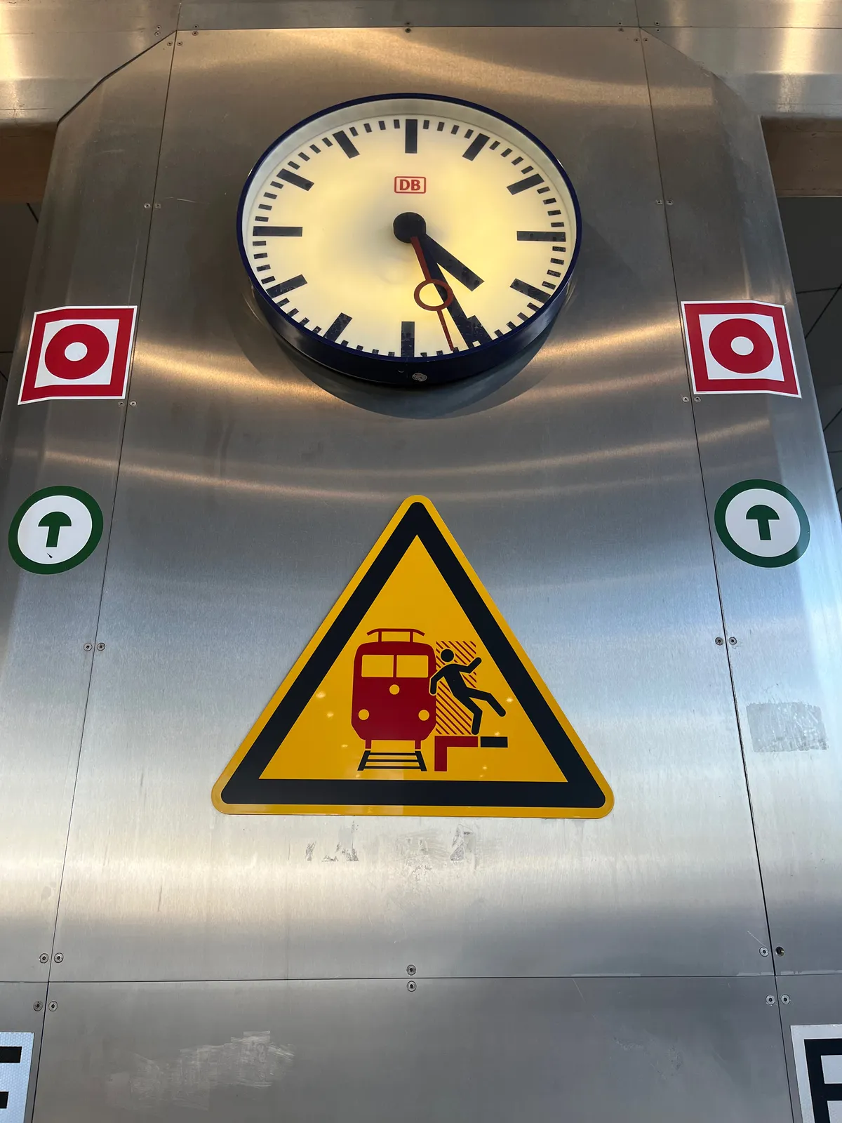 A large warning sticker warns not to fall onto the train tracks. There are a few more stickers whose meaning are unclear. There’s also a clock that points at 26 minutes past 4.