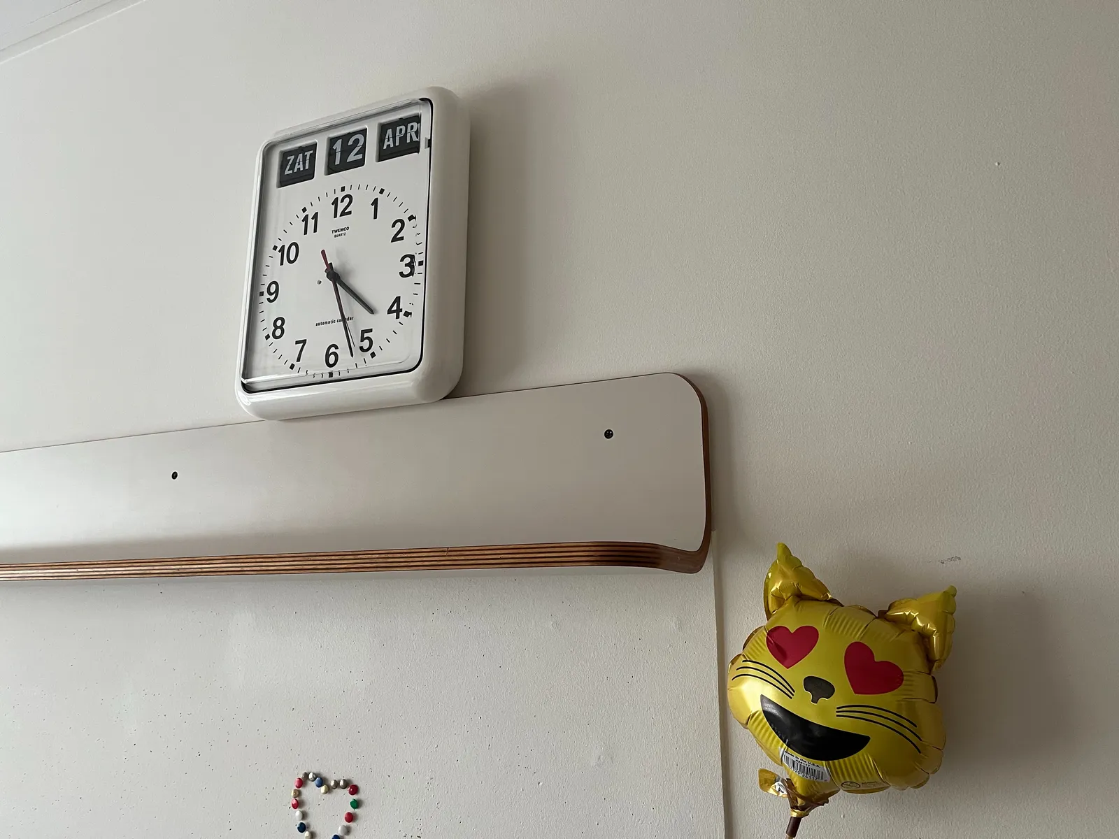 In the foreground a cat shaped balloon with heart shaped eyes. There’s also a simple, heart shaped mosaic on a white board. Above the white board there’s a clock that shows the date, it’s Saturday the 12th of April, and it shows the time as well. It is 27 minutes past 4.