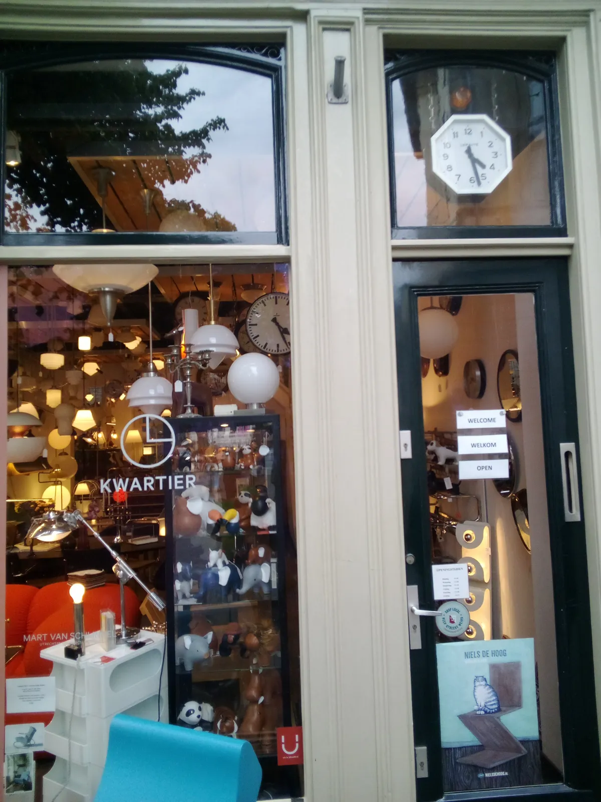 The shopping facade of a design shop. Inside there are lamps and a clock which points at 26 minutes past four. Above the door there’s another clock that points at 27 minutes past four.