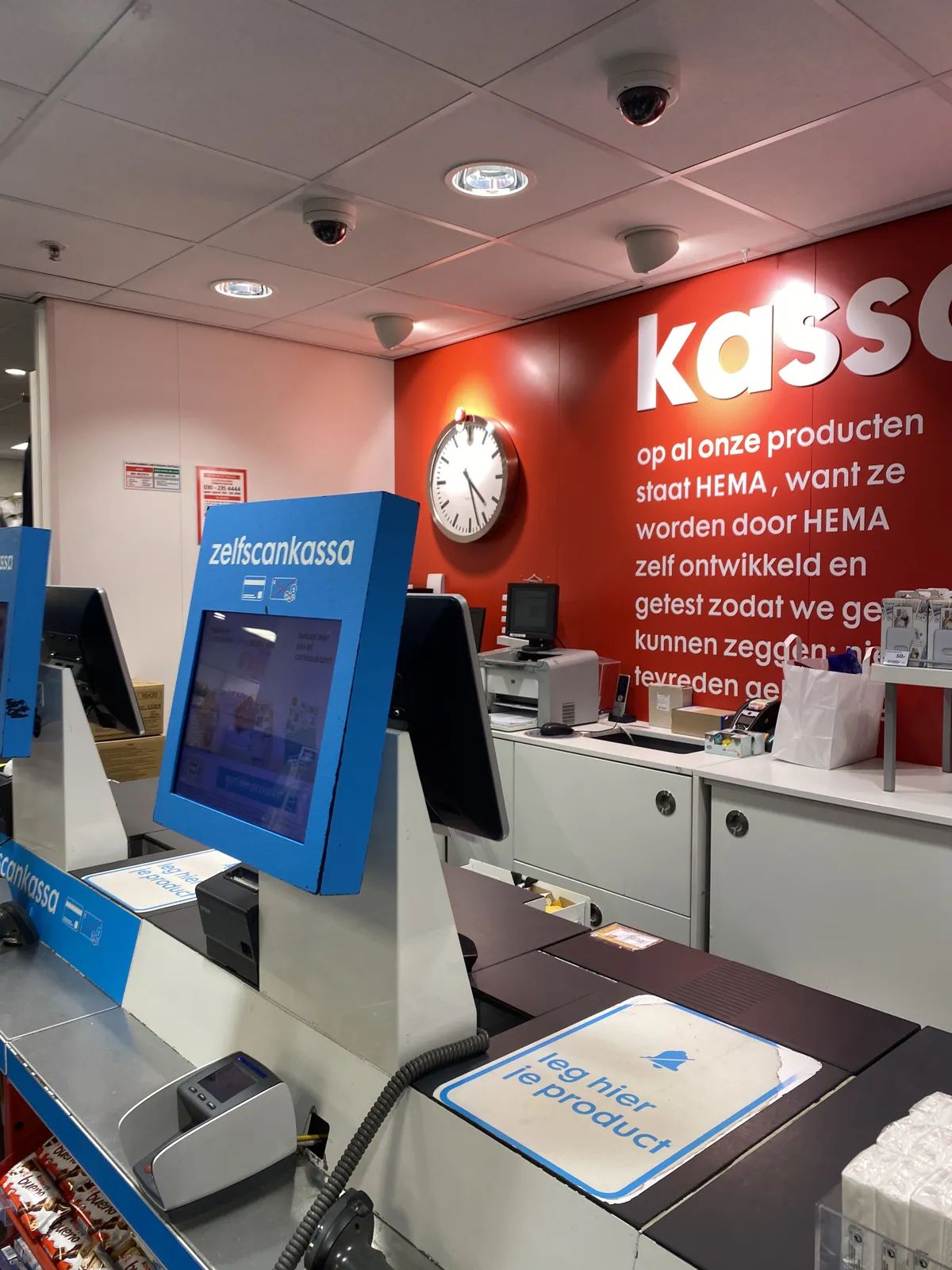 There’s a large clock on a red wall, behind some self-checkout stations in a shop. It points at 27 minutes past 4.