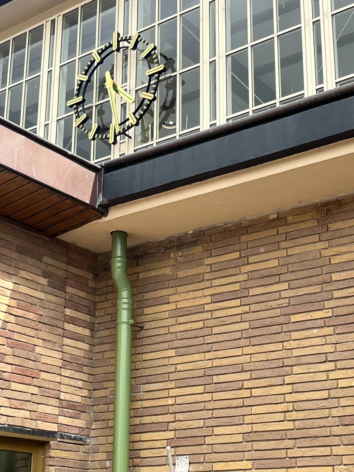 A brick wall with a green drainpipe in the foreground. Behind this wall is the monumental glass facade of the Amstel station in Amsterdam, with the large clock built into it. It points at 29 minutes past 4.