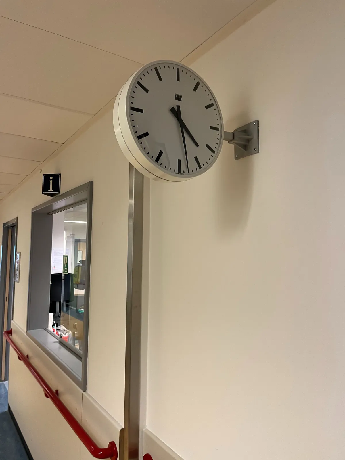 A white, modernist clock with pointy black hands, attached to a white wall, next to a window, in a hallway. It points at 7 minutes past 3.