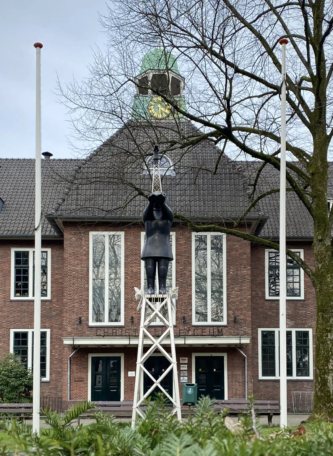 In the middle of the picture there’s a large statue of a person peeking through binoculars. Behind this statue there’s a house with a clock on top of the roof. This clock points at 29 minutes past 4.