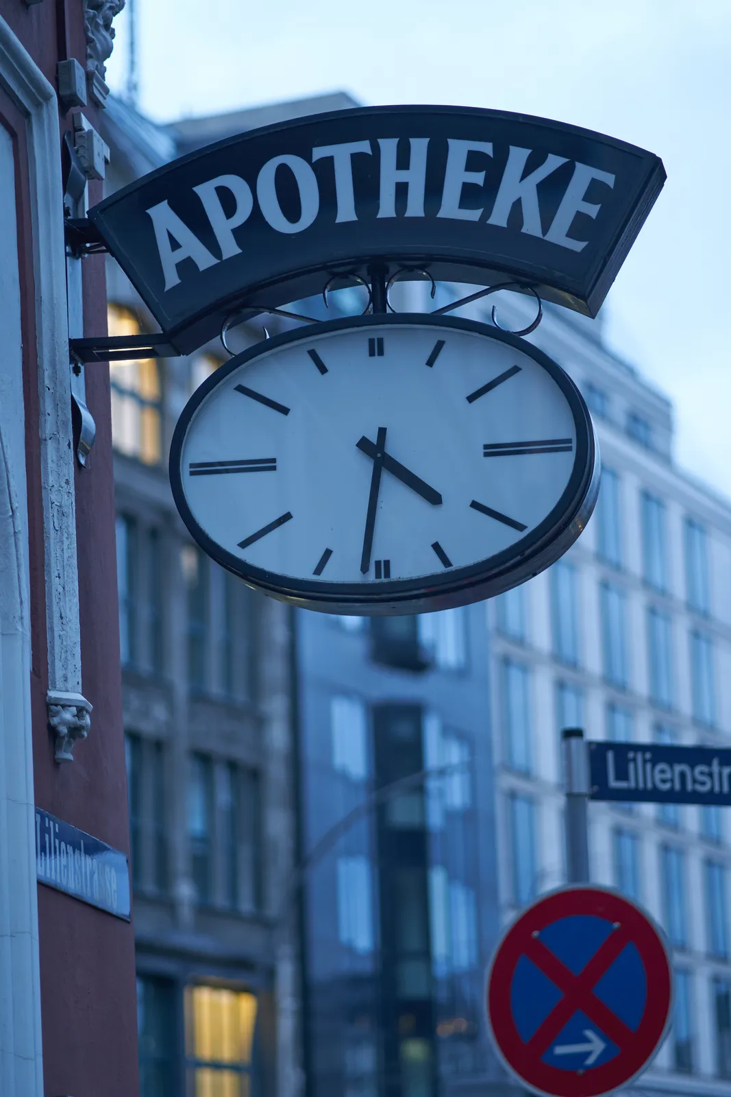 A sign that says Apotheke with an oval clock below it. The clock points at approximately 29 minutes to 5.