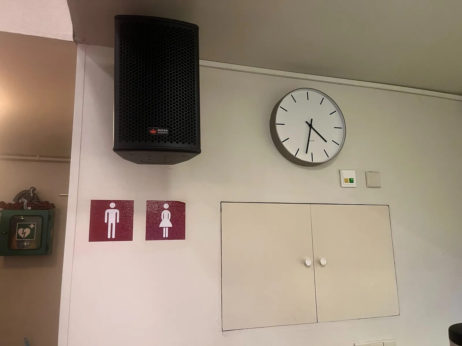 On a white wall, together with a cupboard, a large black speaker and red stickers with a symbol of a man and a woman, there’s a clock. It points at approximately 32 minutes past 4.