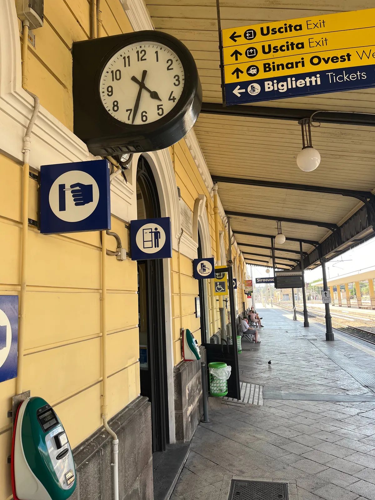 A train station in Italy, in the afternoon in summer. Some signs point at the exits, at the ticket booth, and at Binari Ovest. There’s also a clock which points at 33 minutes past 4.