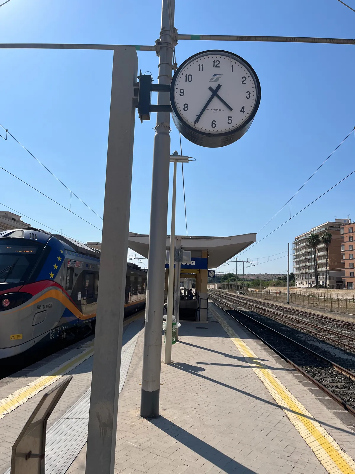 A bright blue, summery sky. On the left there’s a train standing still on the platform. On the right there’s a large appartment building. In the middle there’s a clock that points at 34 minutes past 4.