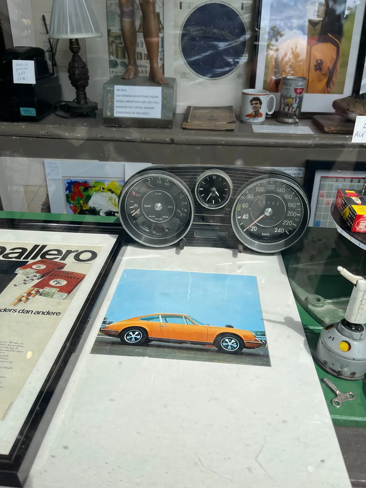 In this shop window of a thrift shop there’s a poster of a yellow sports car, some other posters, the feet of a statue can be seen, and there’s the dashboard of an older car, with a few dials: the one for speed on the right goes up to 240, then there are on the left a few dials for tank, temp and oel. In the middle there’s a smaller clock. It points at 35 minutes past four